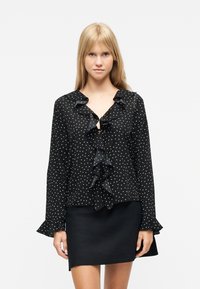 Νεαρή γυναίκα που φοράει μαύρο μπλουζάκι με μακριά μανίκια, λευκές πουά και βολάν, συνδυασμένο με απλή μαύρη φούστα, στέκεται μπροστά σε λευκό φόντο.