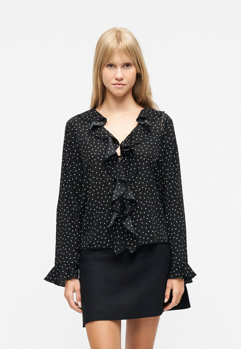 Νεαρή γυναίκα που φοράει μαύρο μπλουζάκι με μακριά μανίκια, λευκές πουά και βολάν, συνδυασμένο με απλή μαύρη φούστα, στέκεται μπροστά σε λευκό φόντο.