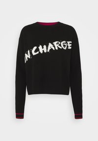 Sweater noir court à manches longues, avec le texte blanc "IN CHARGE", des poignets côtelés avec des accents roses, et un design à col rond.