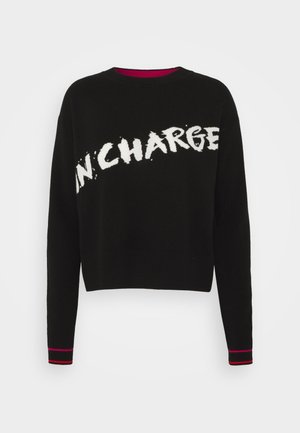 Sweater noir court à manches longues, avec le texte blanc "IN CHARGE", des poignets côtelés avec des accents roses, et un design à col rond.