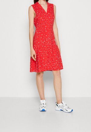 Frau trägt ein ärmelloses rotes Kleid mit kleinem weißem und schwarzem Blumenmuster, kombiniert mit weißen und blauen Sportschuhen.