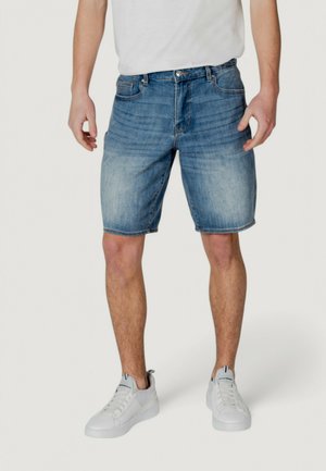 Shorts en denim pour hommes de couleur bleu clair, coupe régulière, avec un délavage, cinq poches et fermeture à bouton en métal, associés à des baskets blanches.