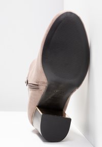 Bottine beige en daim avec semelle noire texturée, fermeture éclair latérale et talon bloc avec accent métallique, vue par le dessous.