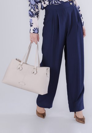 Person iført marineblå højttaljede bukser, blå blomstret bluse og brune sko med hæl, som holder en stor beige håndtaske.