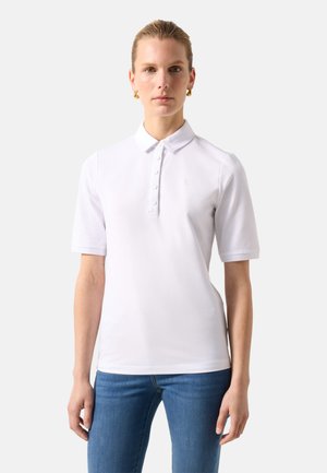 Kvinde iført en hvid kortærmet poloshirt med knapper og blå jeans mod en ensfarvet lys baggrund.