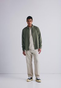 Camicia verde oliva a maniche lunghe sopra una t-shirt beige, abbinata a pantaloni crema e sneaker nere con suole gialle.