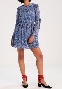 Vestido floral azul con mangas largas transparentes y cintura elástica. Zapatos negros de punta con correas rojas en los tobillos. Tela suave y ligera.