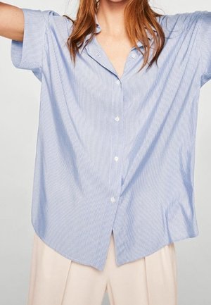Personne portant une chemise ample à rayures bleu clair et blanc avec manches courtes et un pantalon couleur crème, les bras légèrement levés.