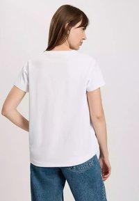 Biała koszulka z krótkim rękawem o luźnym kroju, gładki materiał bawełniany, okrągły dekolt i lekko zaokrąglony dół, noszona z niebieskimi dżinsami.