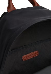 Black backpack interior with a brown leather label stitched inside reading "Tommy Hilfiger" beneath a brown leather handle.