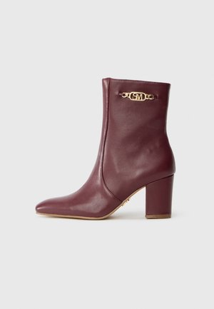Bota de couro burgundy até ao tornozelo com salto bloco e detalhe de corrente dourada, apresentando iniciais gravadas de um lado, sobre fundo branco.