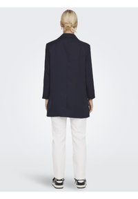 Blazer lungo blu navy con spacco posteriore, chiusura a un bottone e maniche lunghe, abbinato a pantaloni bianchi e sneakers nere.