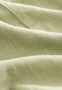 Tissu vert clair avec un tissage texturé, présentant des couches douces et pliées qui mettent en valeur ses fibres naturelles et son éclat subtil.