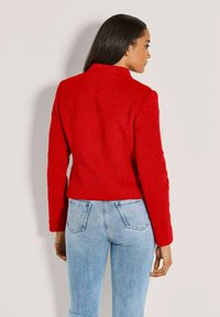 Rote Jacke mit strukturierter Oberfläche und kurzem Schnitt, ausgestattet mit hohem Kragen und einzigartigem Muster. Kombiniert mit hellblauen Jeans.