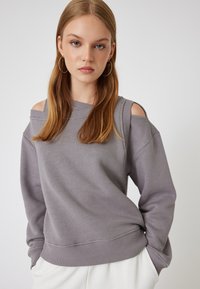 Felpa grigia con spalle scoperte, maniche lunghe, polsini e orlo a coste. Tessuto morbido; vestibilità comoda. Design semplice senza motivi o dettagli.