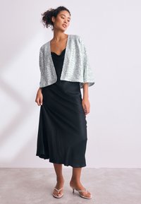 Schwarzes Satinkleid mit Wasserfallausschnitt, kombiniert mit einer silbernen, mit Pailletten besetzten Bolerojacke. Dazu klare Sandalen mit Absatz. Glatte Textur.