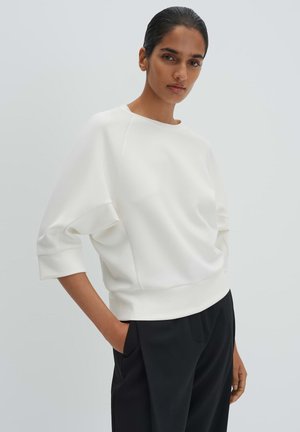 Frau mit dunklen Haaren, die eine weiße Dreiviertelarmbluse und schwarze Hosen trägt, steht mit einer Hand in der Tasche vor einem schlichten hellen Hintergrund.