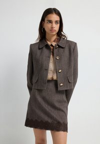 Vrouw in een bruin, gestructureerd, cropped jack met gouden knopen en een bijpassende rok met kantafwerking, poserend tegen een effen lichte achtergrond.