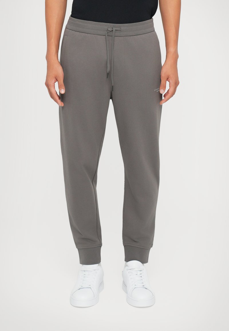Grijze joggingsbroek met elastische tailleband en trekkoord, voorzien van zijzakken en geribbelde boorden. Gecombineerd met witte sneakers.