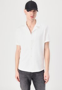 Chemise blanche à manches courtes avec col, présentant de légers boutons et une coupe ample, associée à un jean gris foncé et une casquette noire.