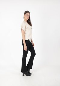 Top beige de manga corta con escote en V y detalle de botones en el frente, combinado con pantalones acampanados negros y botas negras de tobillo, de pie sobre un fondo blanco.