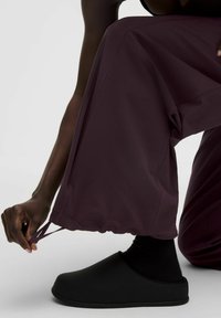 Osoba v tmavo purpurových nohaviciach a čiernych klzákov prilieha elastický kábel na okraji nohavíc, keď sa schováva na bielom podlahe.