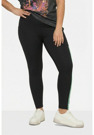 Mallas con franjas laterales verdes combinadas con zapatillas blancas y una camiseta gráfica negra, modelo de pie con una mano en la cadera.