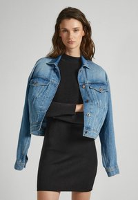 Giacca in denim azzurro chiaro con due tasche sul petto, chiusura a bottoni e vestibilità corta. Indossata sopra un vestito nero a coste con maniche lunghe.