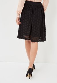 Falda negra hasta la rodilla con encaje texturizado de patrón de diamantes, con una cintura entallada. Combinada con zapatos de tacón alto negros.