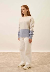 Brede trui van crème en lichtblauw, met een geribbelde structuur, brede manchetten en een ontspannen pasvorm, gecombineerd met witte wijde broekspijpen.
