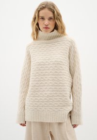 Texturierter cremefarbener Rollkragenpullover mit wellenförmigem Muster und gerippten Bündchen. Hergestellt aus einem weichen Material, mit lockerer Passform und seitlichen Schlitzen.