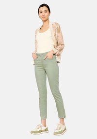 Lichte roze bloemen bomberjack over een crèmekleurig tanktop, gecombineerd met lichtgroene slim-fit broek en beige-groene sneakers.