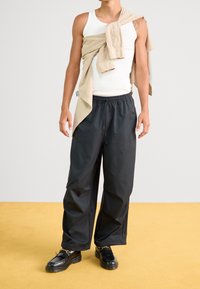 Schwarzweite, lockere Hose mit elastischem Bund, weißes Tanktop und ein beigefarbenes, knöpfbares Hemd über die Schultern gelegt. Schwarze Schuhe.