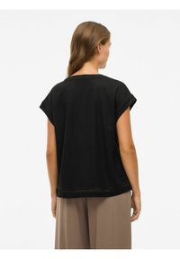 Frau mit langen braunen Haaren, die ein lockeres schwarzes Top mit kurzen Ärmeln und beige weit geschnittene Hose trägt, von hinten vor einem weißen Hintergrund betrachtet.