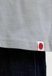 Hellgraues Baumwoll-T-Shirt mit einem roten Punkt-Label am Saum, kombiniert mit dunkelblauer Jeanshose. Nahaufnahme zeigt die Nahtdetails.
