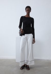 Svart langermet topp med knutdetalj, kombinert med hvite bukser med vide ben og svart piping. Modellen bruker en liten crossbody-veske og hælsandaler.