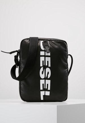 Sac bandoulière rectangulaire noir avec logo Diesel blanc, sangle réglable et fermeture éclair, posé sur une surface blanche.