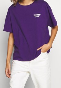 Lila bomullströja med korta ärmar och rund halsringning. Har vit broderad text på vänster bröstområde. Matchas med vita byxor.