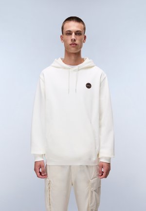 Ung mand iført en hvid hættetrøje med en lille rund logo på brystet og lysebeige cargobukser.