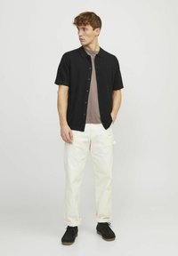 Cămașă neagră cu mâneci scurte și nasturi peste un tricou marron, asortată cu pantaloni crem răsuciți și pantofi negri, având un design casual.