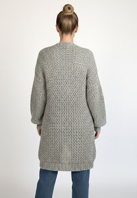 Długi szary dziergany kardigan z fakturowanym wzorem, szerokimi rękawami i ściągaczem u dołu, w połączeniu z niebieskimi spodniami.