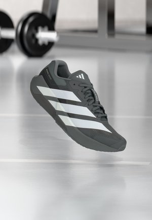 Chaussure de sport gris foncé avec des bandes blanches flottant à l'intérieur sur une surface réfléchissante, équipement de gym flou en arrière-plan.