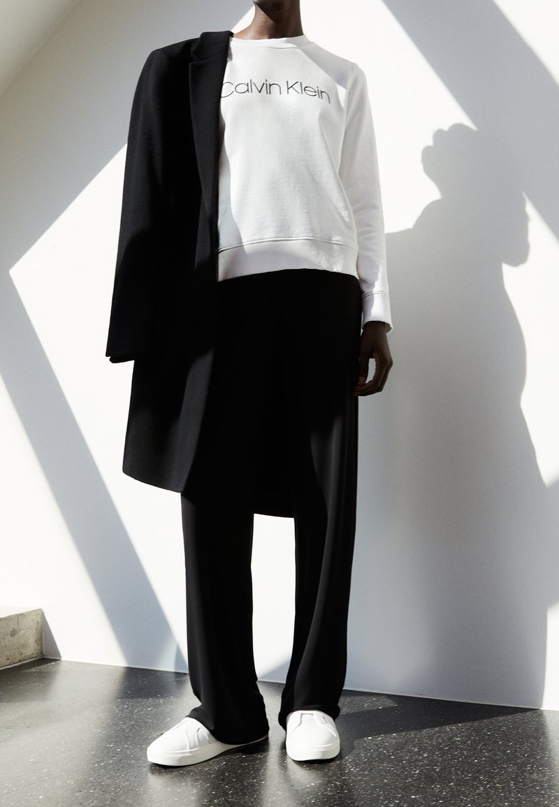 Felpa bianca con logo "Calvin Klein", cappotto nero appoggiato sulla spalla, pantaloni neri larghi e scarpe bianche slip-on, su uno sfondo luminoso.