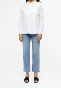 Camisa blanca de manga larga y botones combinada con jeans rectos de color azul claro y sandalias negras de tipo slides. Diseño simple y limpio, adecuado para un estilo casual.