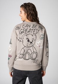 Young woman with curly hair smiles, wearing a gray sweatshirt featuring a teddy bear drawing and text "You can be the greatest" on the back.