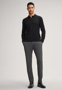 Schwarzer Pullover mit einem Halbreißverschlusskragen, kombiniert mit grauen strukturierten Hosen. Das Model steht mit den Händen in den Taschen und trägt schwarze Lederschuhe.