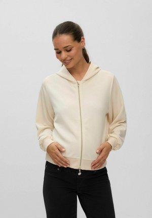Femme portant un sweat à capuche crème zippé et un pantalon noir, souriante, les mains posées délicatement sur le bas de son ventre sur un fond uni.