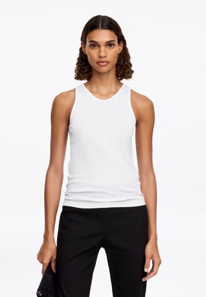Femme aux cheveux bouclés portant un haut blanc sans manches et un pantalon noir, tenant une pochette noire sur un fond blanc uni.