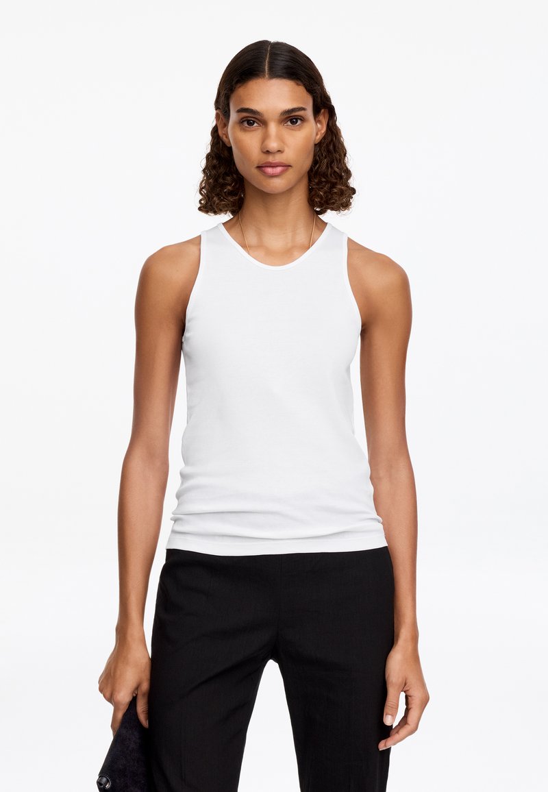 Femme aux cheveux bouclés portant un haut blanc sans manches et un pantalon noir, tenant une pochette noire sur un fond blanc uni.