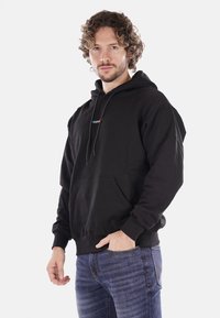 Felpa nera in cotone con tasca frontale, cappuccio con cordino e logo "PROPAGANDA" multicolore sul petto, indossata con jeans blu.